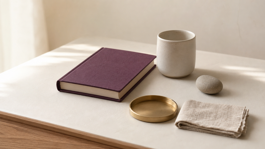 Plum journal, ceramic cup, smooth stone, gold tray, and folded linen on a cream tabletop.