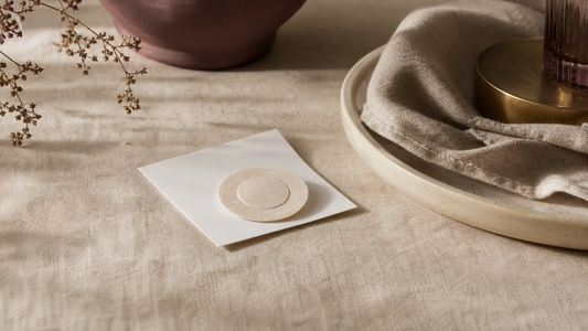 Small unbranded round adhesive disc on paper backing beside a ceramic tray and folded neutral cloth.